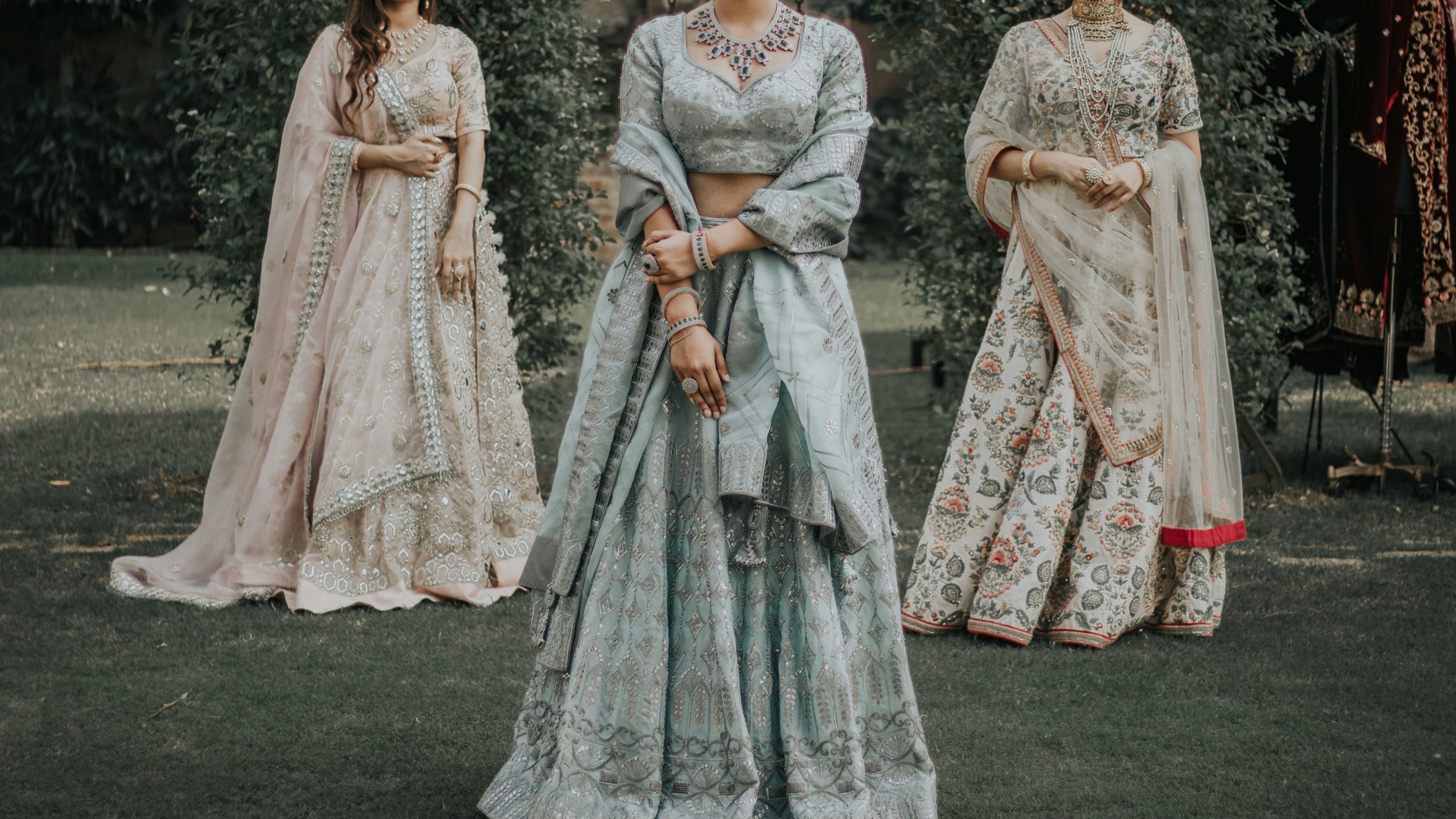 Three women in traditional embroidered dresses standing outdoors with greenery in the background.